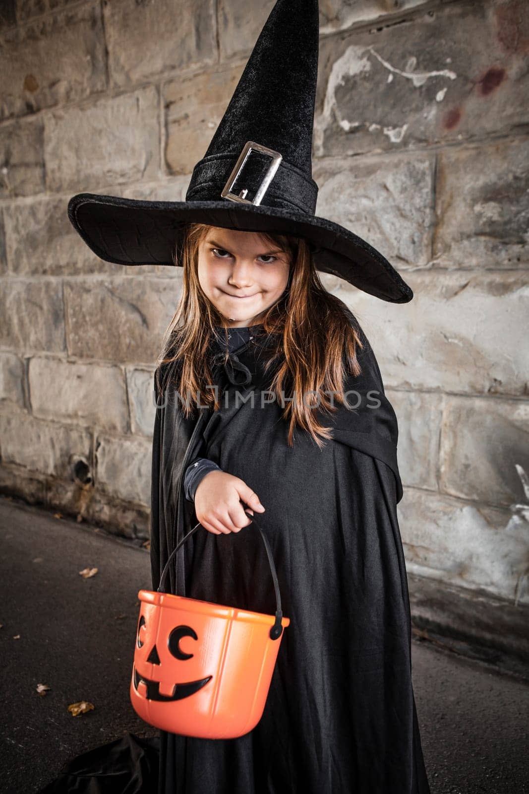Young girl dressed in a witch costume with tall black hat and holding a pumpkin pail for halloween.