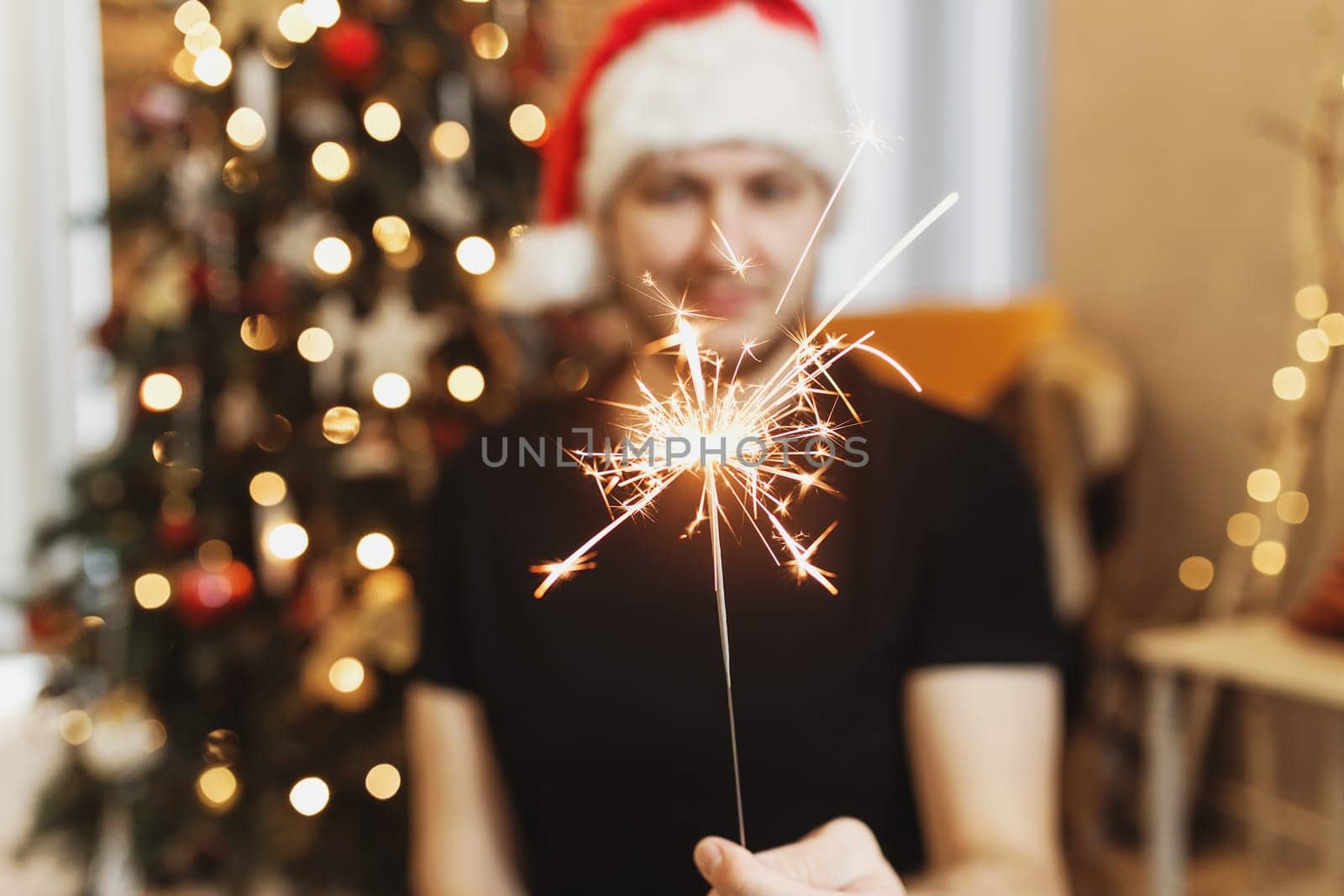 Smiling man in red santa hat or cap holding burning Christmas or New Year sparkler lights in hand. Christmas fir tree with garland lights on blurred background. Festive mood. High quality photo
