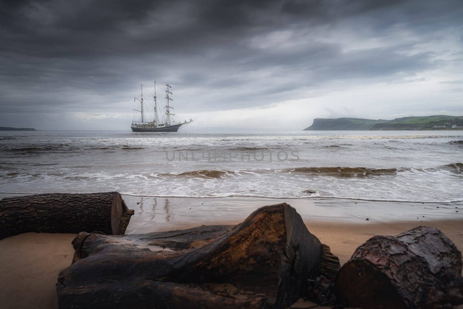 Dramatic storm sky rolling over anchored tall ship near Northern Ireland coast, Fair Head in far distance, seen from behind of drift wood on a beach