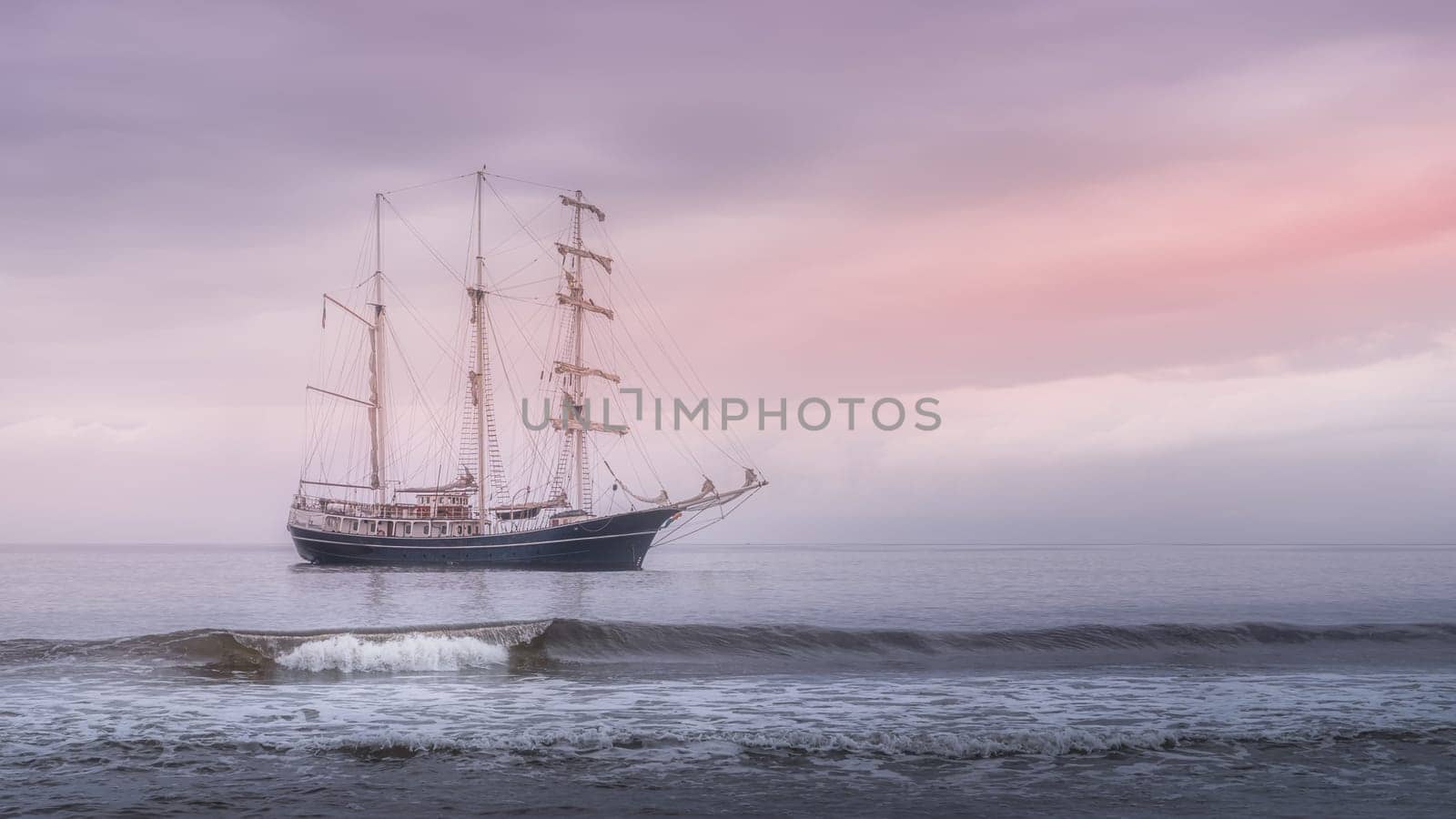 Rolling wave in front of beautiful tall ship covered in haze with soft morning sky, anchored near Northern Ireland coast, Ballycastle