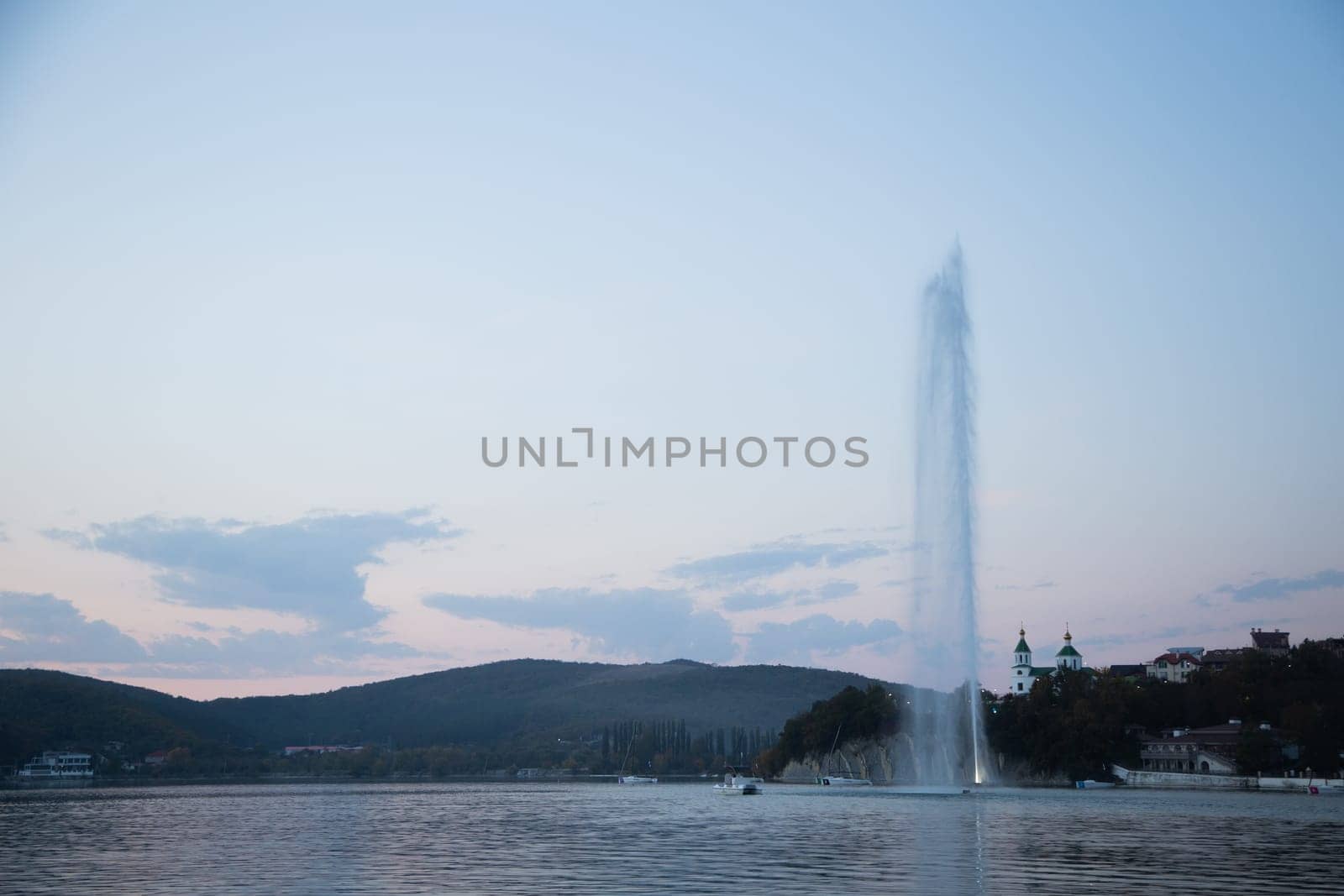 A fountain several meters high spouts from the center of the lake, you can see the mountains and the church, evening. Abrau Durso, Krasnodar Territory. High quality photo