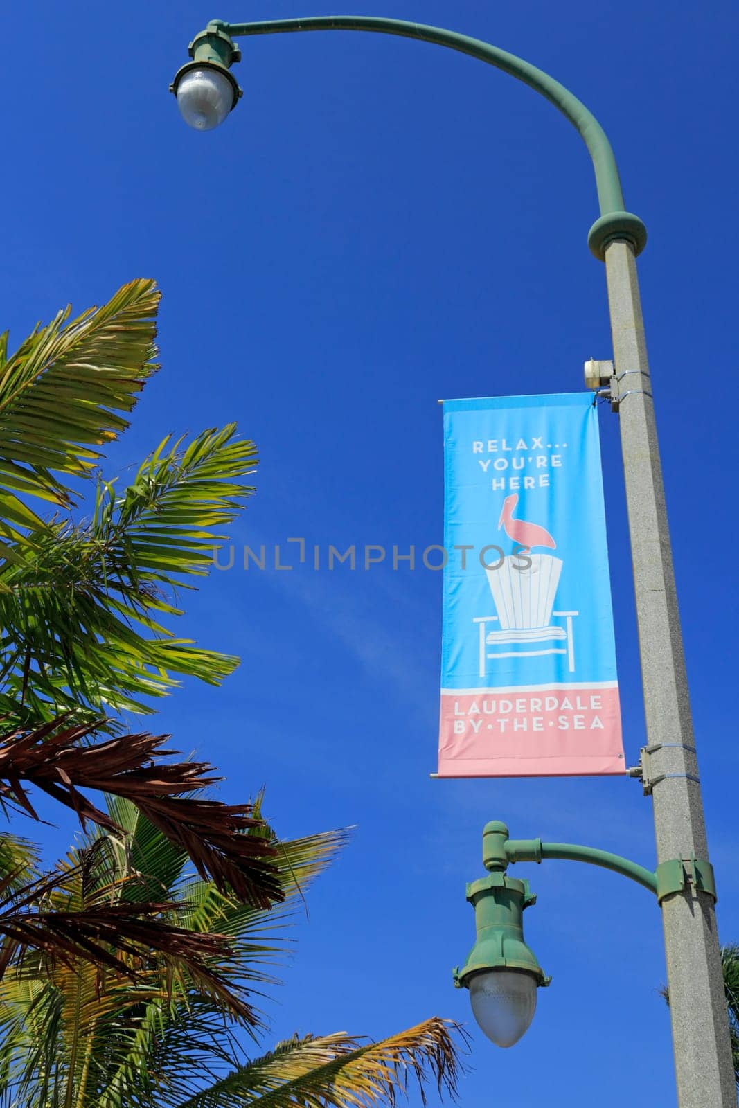 LAUDERDALE-BY-THE-SEA, FL, USA - APRIL 7, 2014: A red white and blue sign that says, Relax…You’re Here, includes a pelican bird and chair hanging from a lamp post near palm tree fronds. A street sign in a tropical community that says, Relax…You’re Here, LAUDERDALE-BY-THE-SEA.