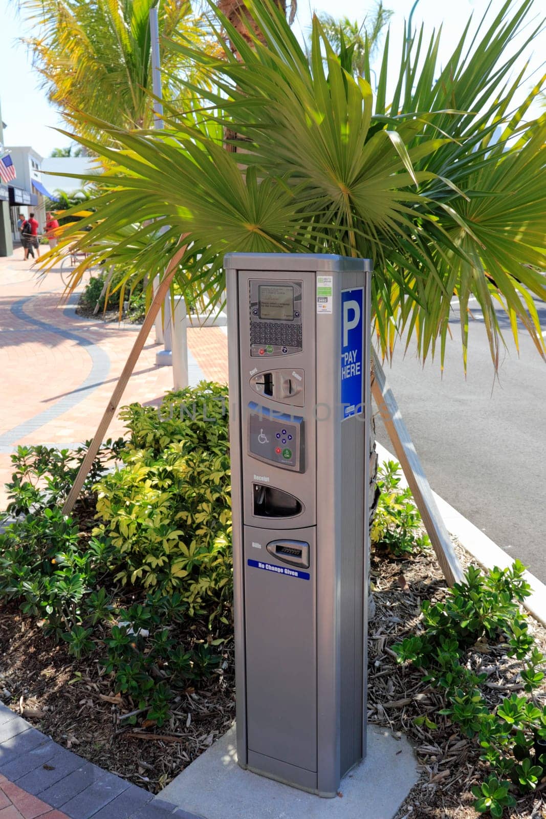 LAUDERDALE-BY-THE-SEA, FL, USA - APRIL 7, 2014: Closeup of an electric parking meter with signage in the shade of palm trees and plants of a newly redesigned parking lot. New silver gray parking meter with digital display panels in a shady area of a newer walkway and parking lot buildings and a few people on the sidewalk.