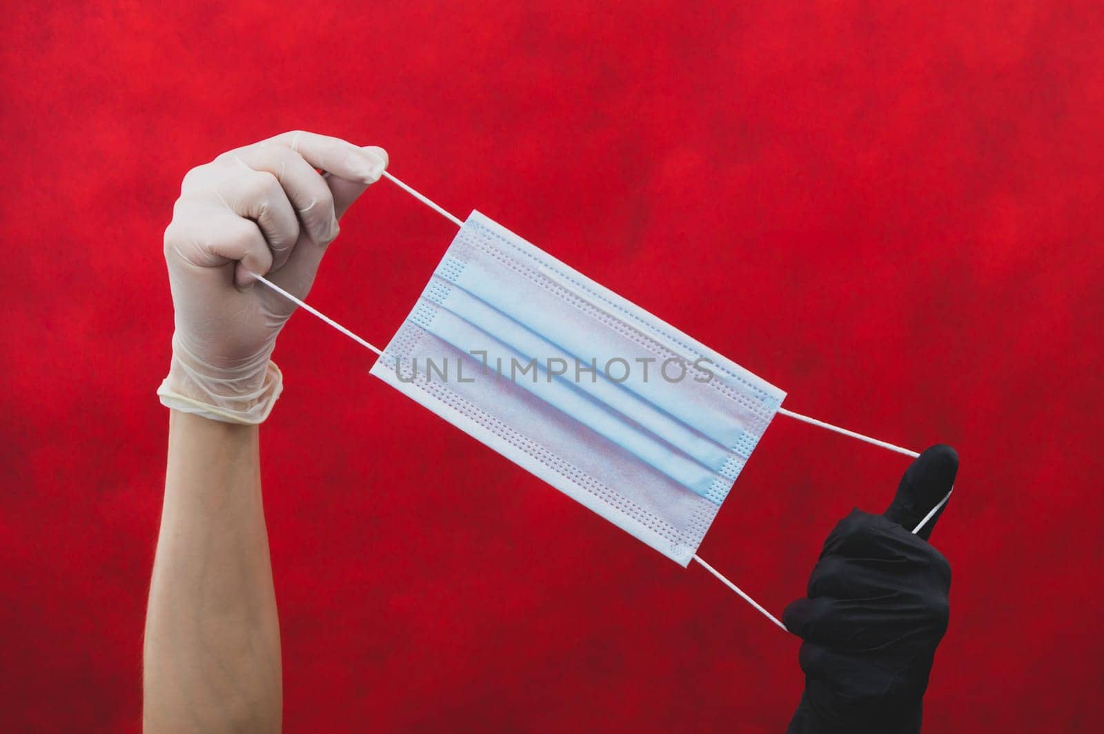 Hands in different protective gloves stretch a medical mask on a red background. one hand in black glove, the other in white. preventive measures against respiratory illness and virus