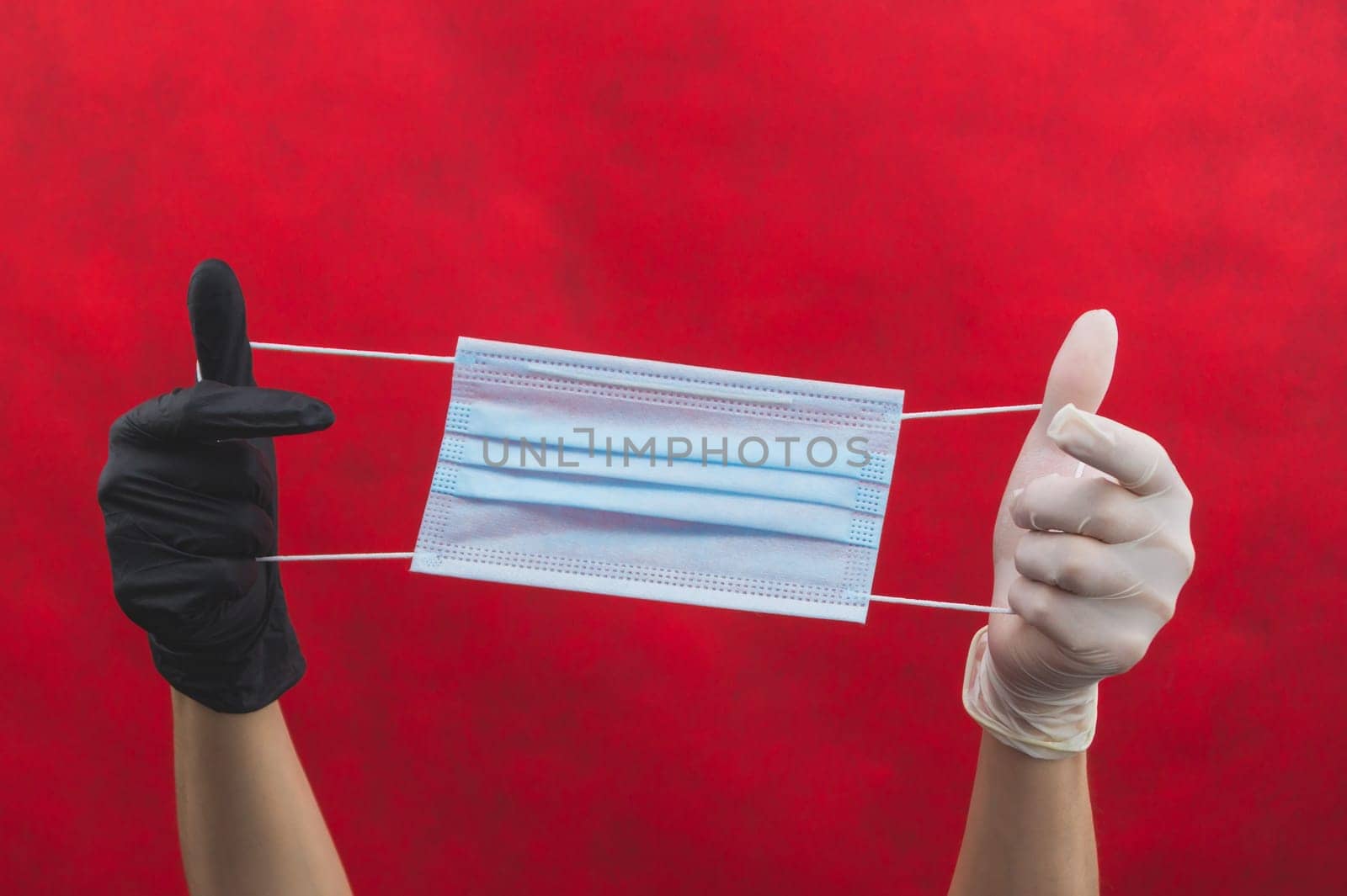 Hands in different protective gloves stretch a medical mask on a red background. one hand in black glove, the other in white. preventive measures against respiratory illness and virus