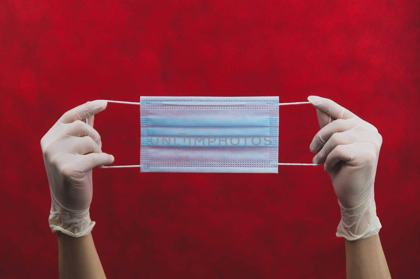 Blue medical mask in hands with white gloves on a red background. preventive measures against respiratory illness and virus. hands in white surgical gloves holds disposable mask for protection