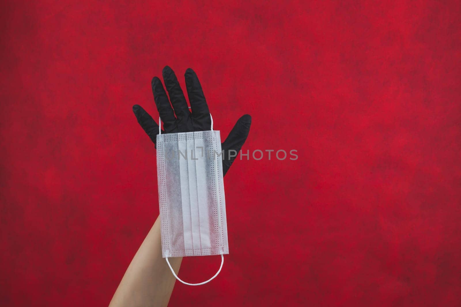 White surgical mask hangs on the hand with a black medical glove. preventive measures against respiratory illness and virus. disposable mask for protection. red background with copy space
