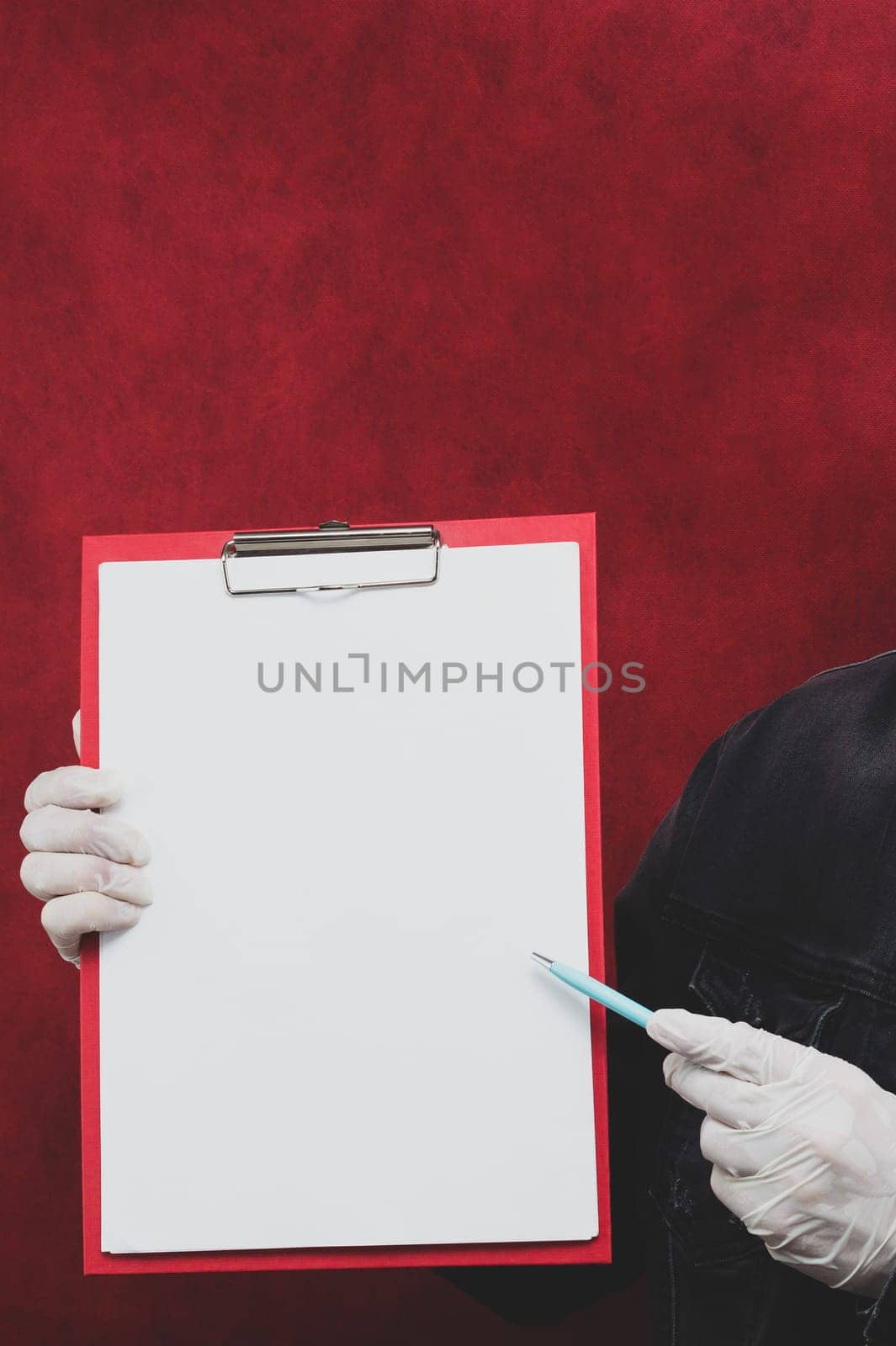 Hand in protective glove holding white blank paper sheet on red background. hand holds a pen and points out the sheet. template mock up. surface of leaflet. copy space
