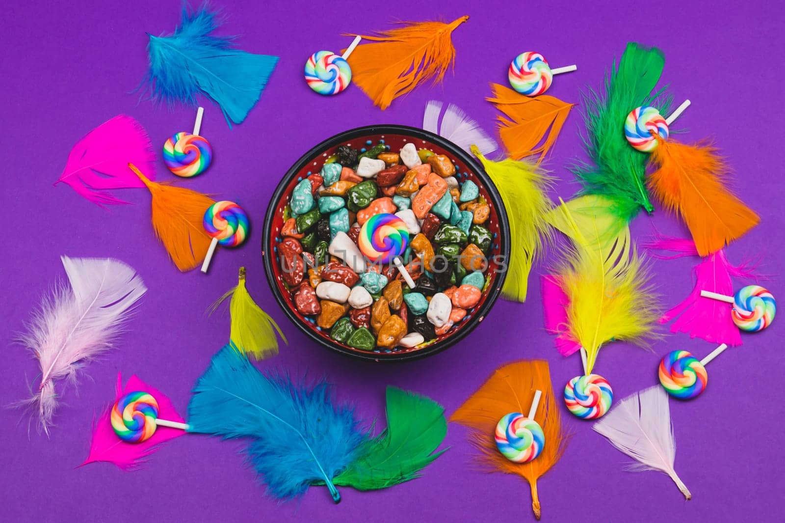 Candy pebbles in a bowl and lollipops. spiral candies and sweets in the form of colored stones. colorful feathers. festive background