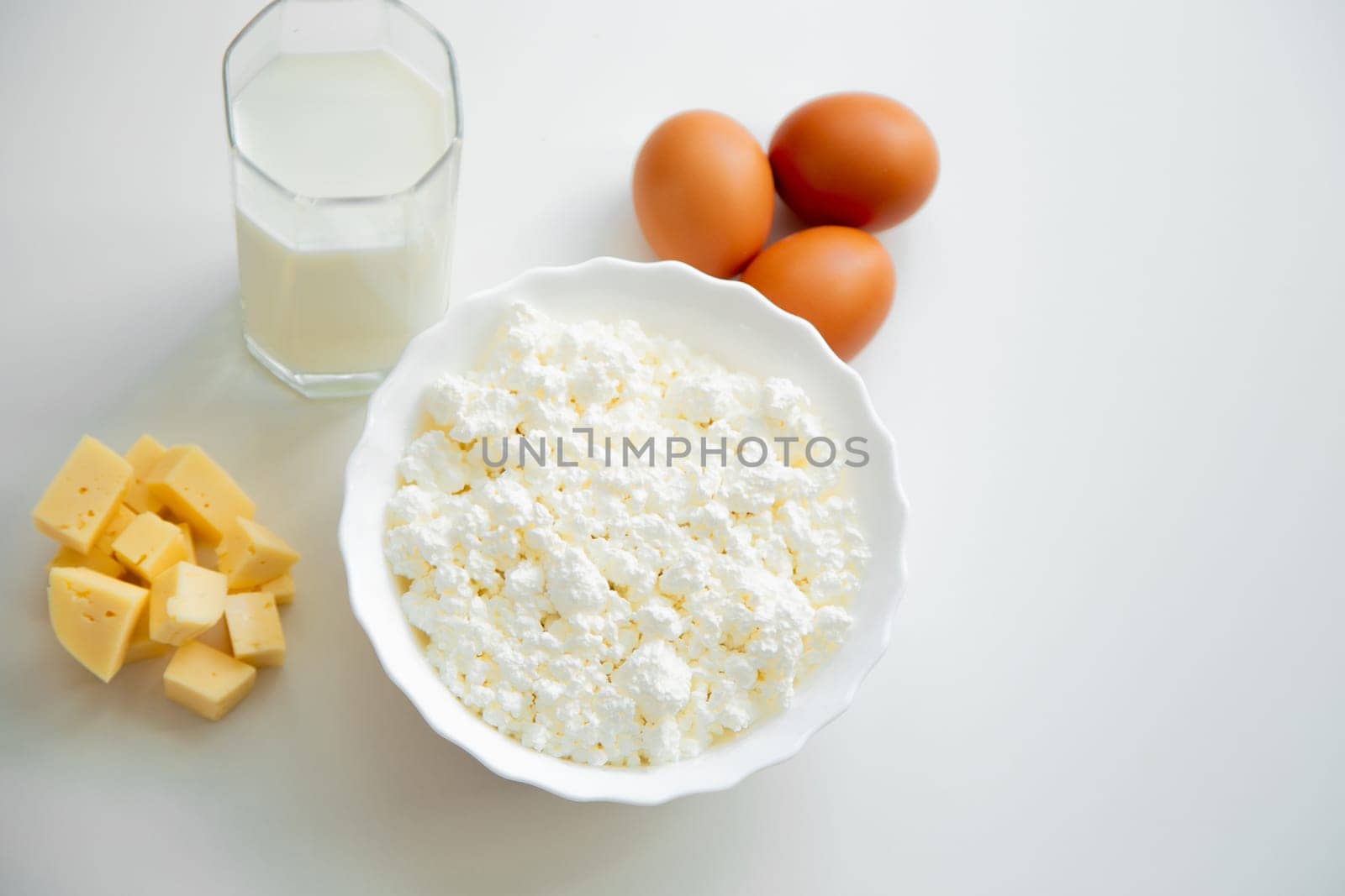 A white bowl with cottage cheese, eggs, diced cheese and a glass of milk stand on a white background. . High quality photo