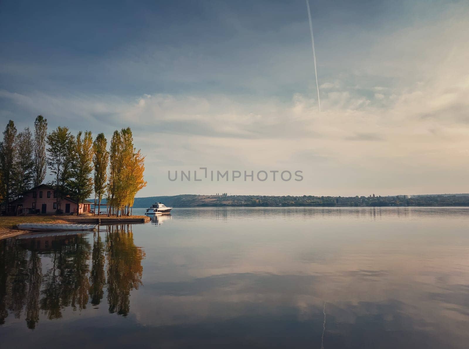 Scenic view with a house on the riverbank of Nistru river and a yacht floating. Autumn season landscape, colorful trees and beautiful sky reflected on the water surface.