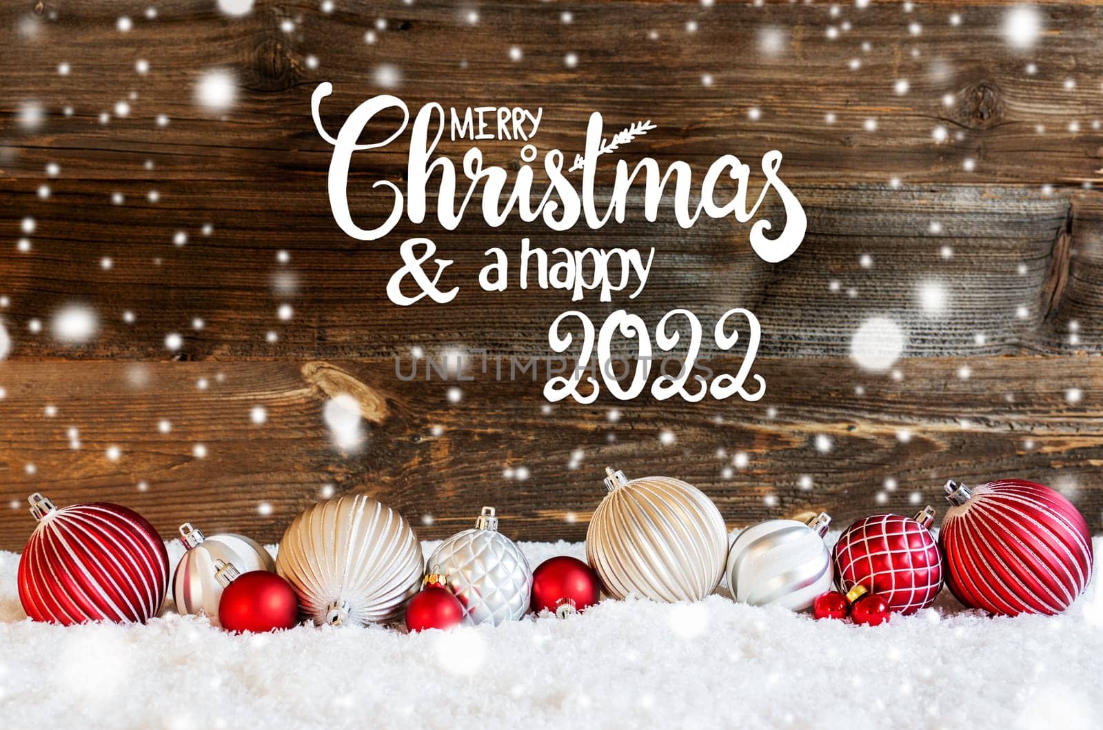 English Calligraphy Merry Christmas And A Happy 2022 On Rustic Brown Wooden Backgroud. Red And White Festive Christmas Ball Ornament On Snow And Snowflakes