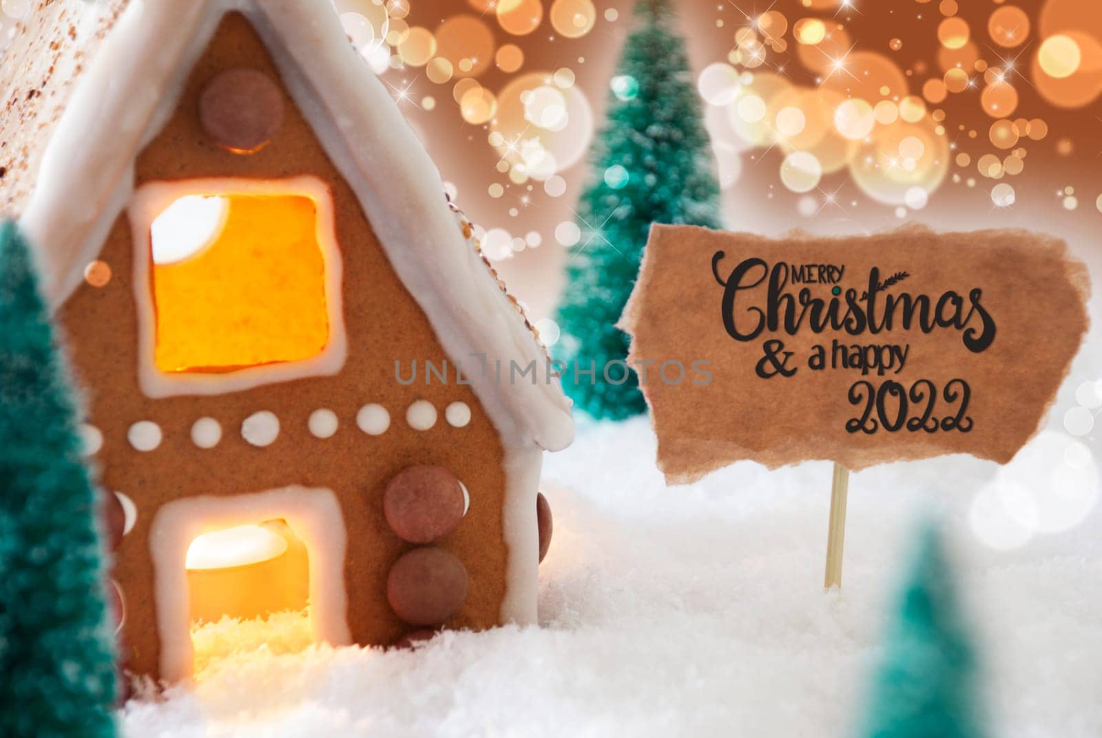 Sign With English Calligraphy Merry Christmas And A Happy 2022. Gingerbread House With Christmas Tree And Snow And Golden Sparkling Background