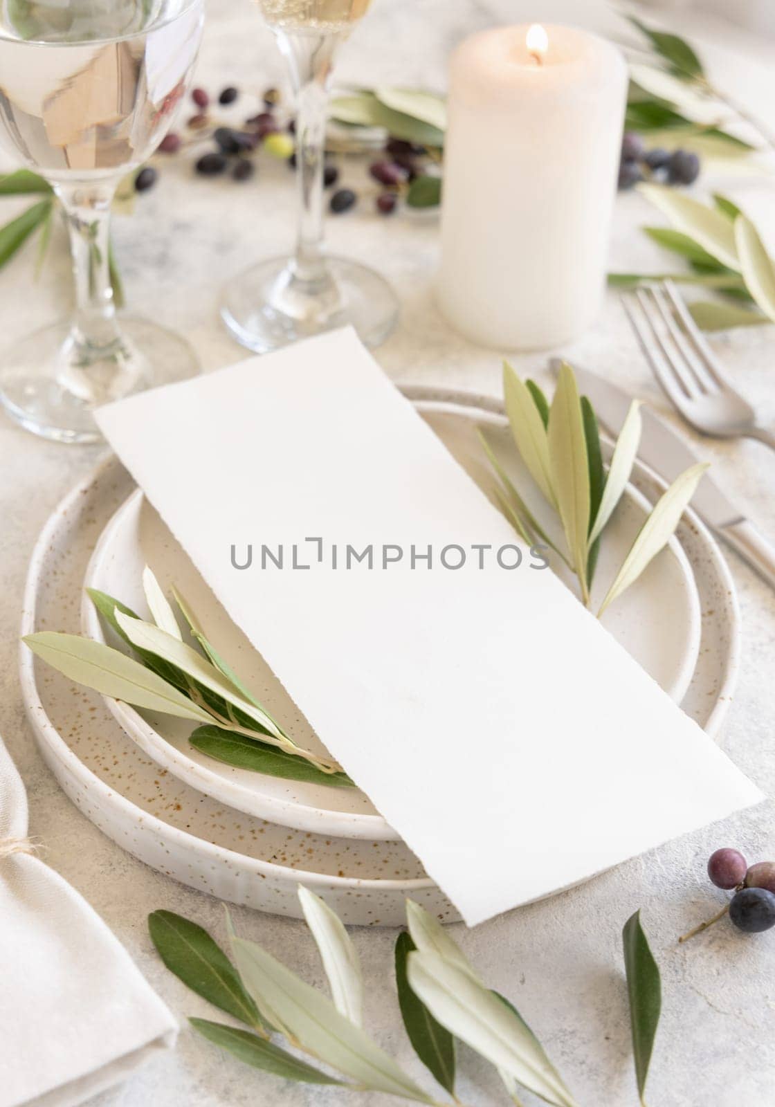 Wedding Table place with a menu card and porcelain plates decorated with olive branches close up. Elegant modern template with vertical blank paper card. Mediterranean flat lay mockup, copy space