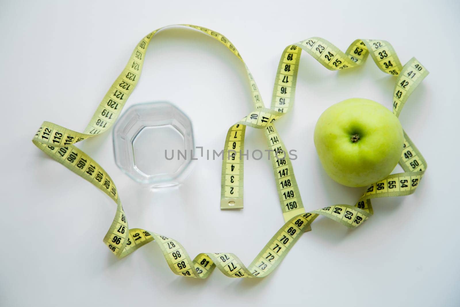 A green apple and a glass of water stand on a white background, they are entwined with a measuring tape. High quality photo