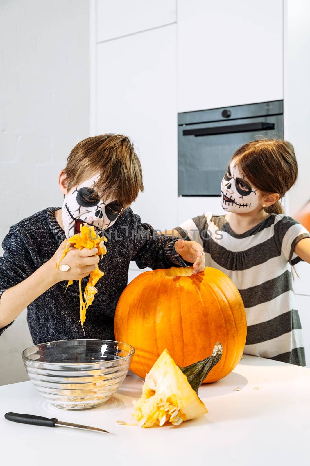 Two young children with skull face paint cutting a pumpkin in their kitchen.