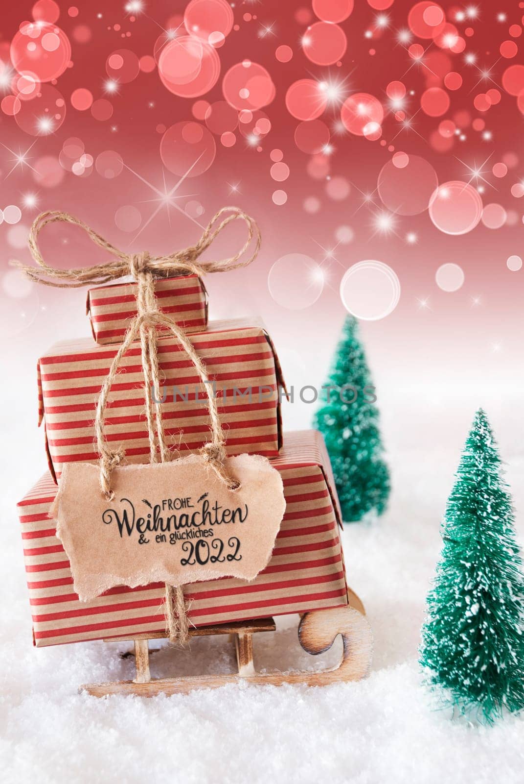 Sled With Christmas Gift And Label With German Frohe Weihnachten Und Ein Glueckliches 2022 Means Merry Christmas And A Happy 2022. Christmas Tree With Snow And Red Sparkling Background