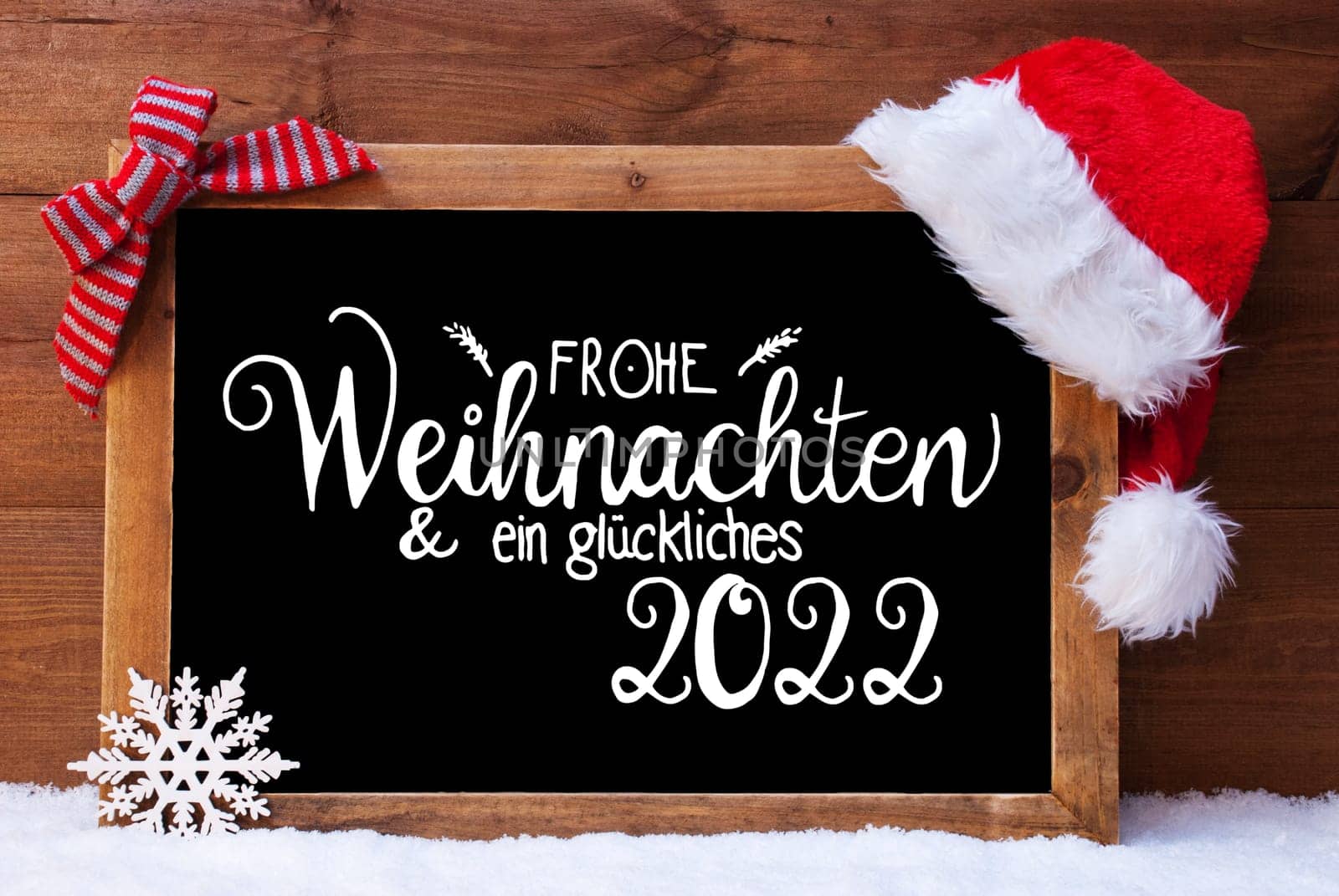 Chalkboard With German Calligraphy Frohe Weihnachten Und Ein Glueckliches 2022 Means Merry Christmas And A Happy 2022. Christmas Decoration Like Santa Hat And Bow. Wooden Background With Snow