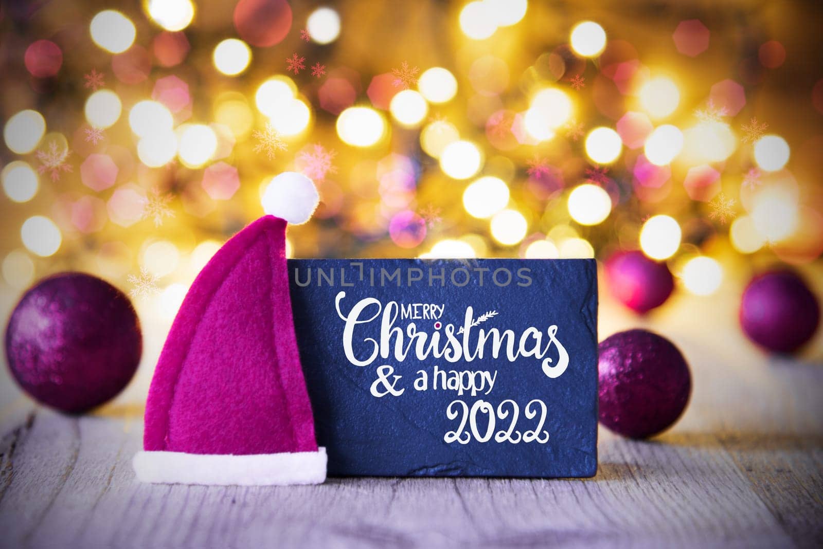 Sighn Wth English Calligraphy Merry Christmas And Happy 2022. Purple Christmas Ball Ornament With Santa Hat. Sparkling Lights Background