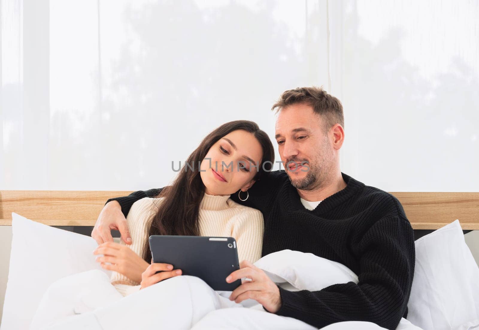 Happy young couple in sweater using a tablet PC while lying in bed at home. Lover relaxing and watching a movie together