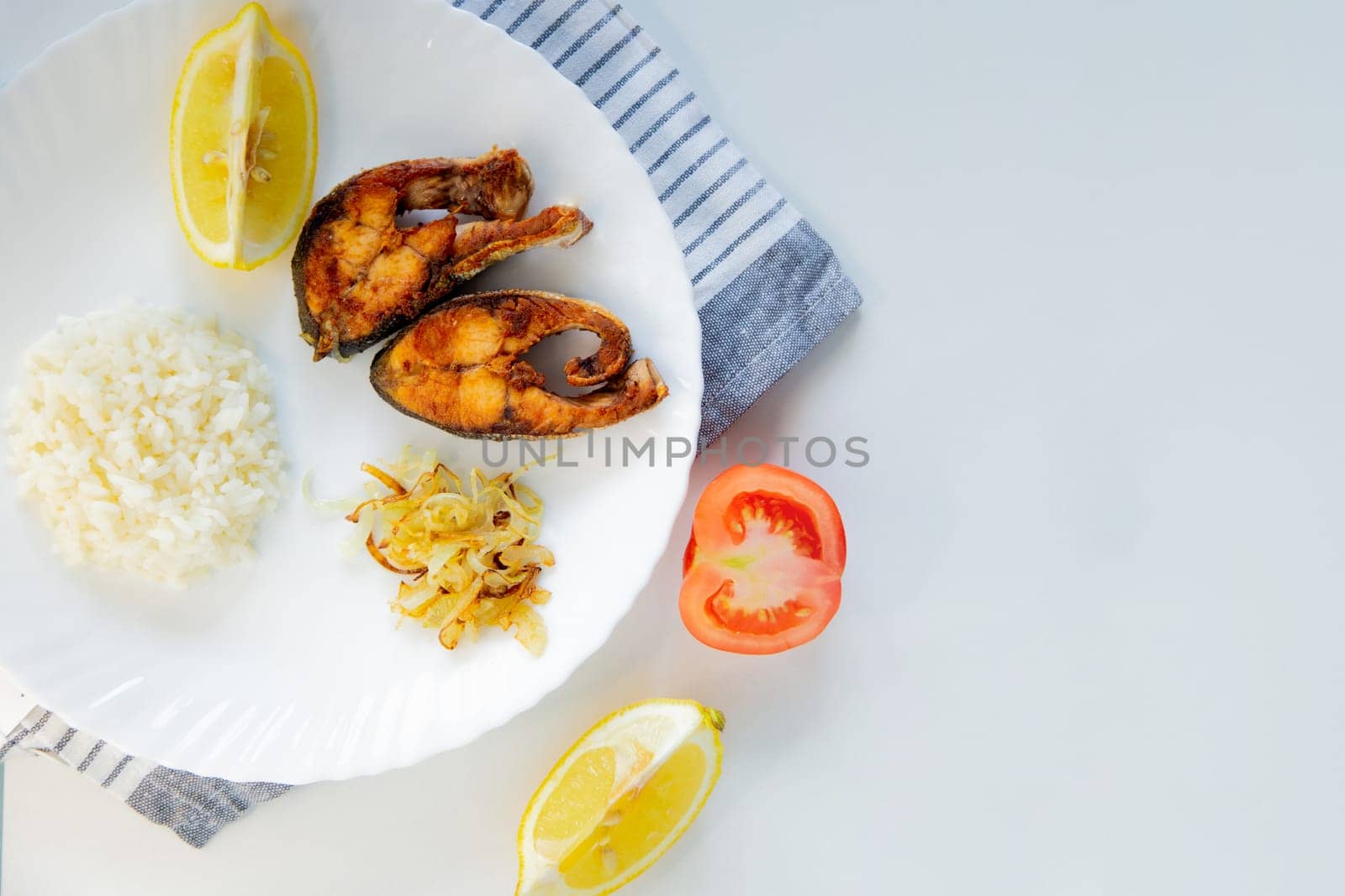 Fried fish with onions, rice and lemon lies on a plate, on a waffle towel and cutlery on a white background. High quality photo