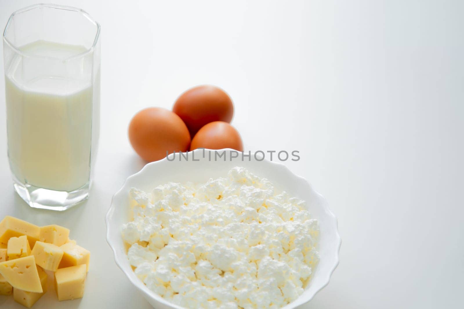 A white bowl with cottage cheese, eggs, diced cheese and a glass of milk stand on a white background. . High quality photo