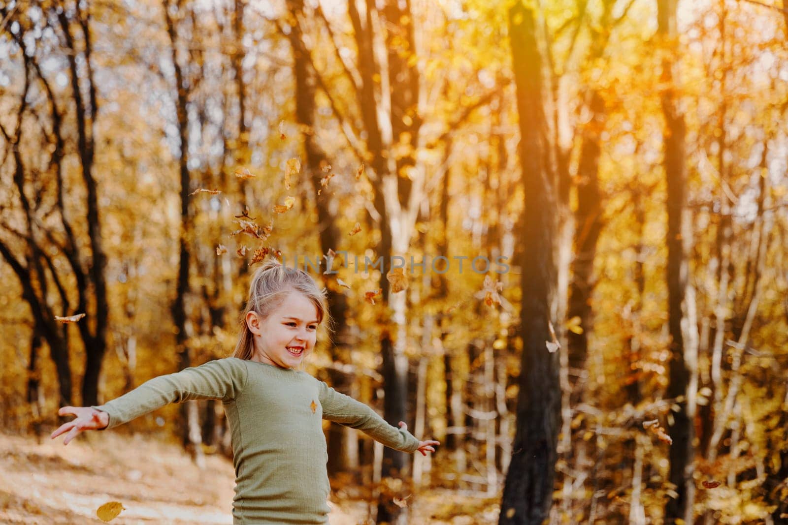 Small happy smiling little caucasian child girl playing with yellow leaves in autumn forest or garden around trees. Kid throws leaves up and they are falling down. Colorful autumn, gold colors around