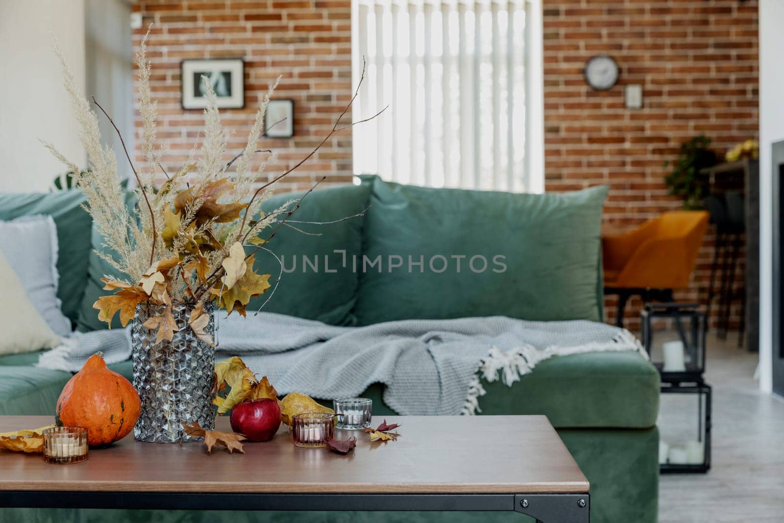 Autumn time decorated home interior. Yellow flowers bouquet in glass vase on wooden stylish table, pumpkin, red apples, leaves, candles, blanket. Green sofa, brick wall and fireplace on background