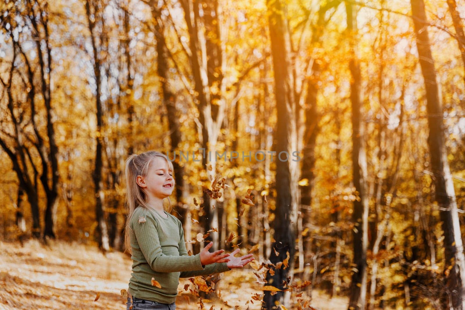 Small happy smiling little caucasian child girl playing with yellow leaves in autumn forest or garden around trees. Kid throws leaves up and they are falling down. Colorful autumn, gold colors around
