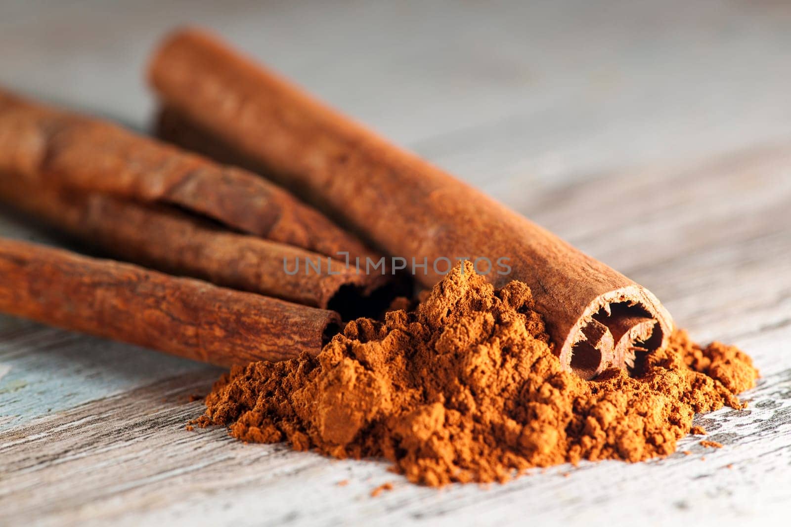 Close-up of several rolled cinnamon sticks with cinnamon powder lying in front of them on a wooden surface with paint residues.
