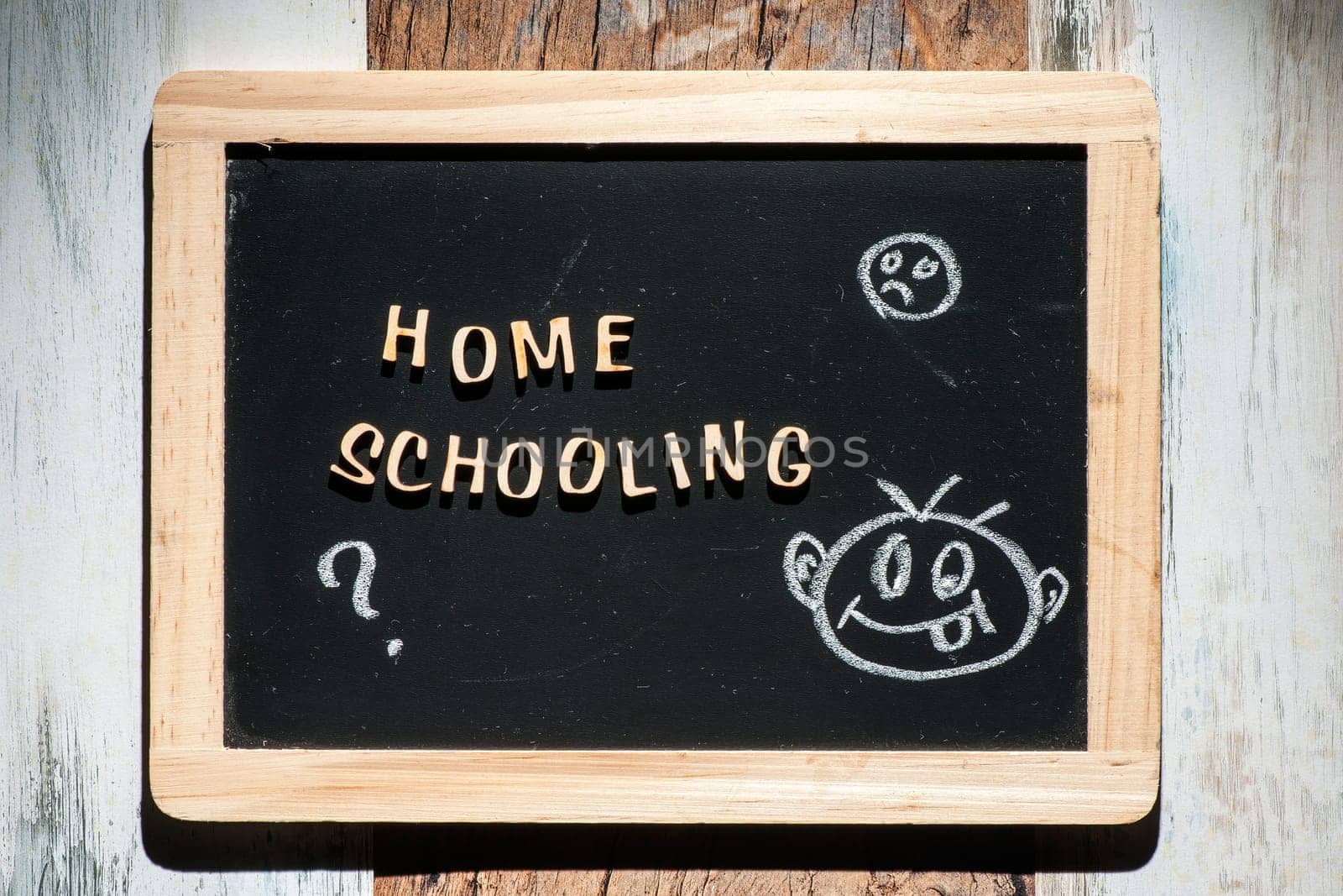 Frontal close-up of a small black chalk board made of wooden letters with the words Home Schooling and a stick figure drawing made of chalk on a wooden surface in the harsh sidelight.