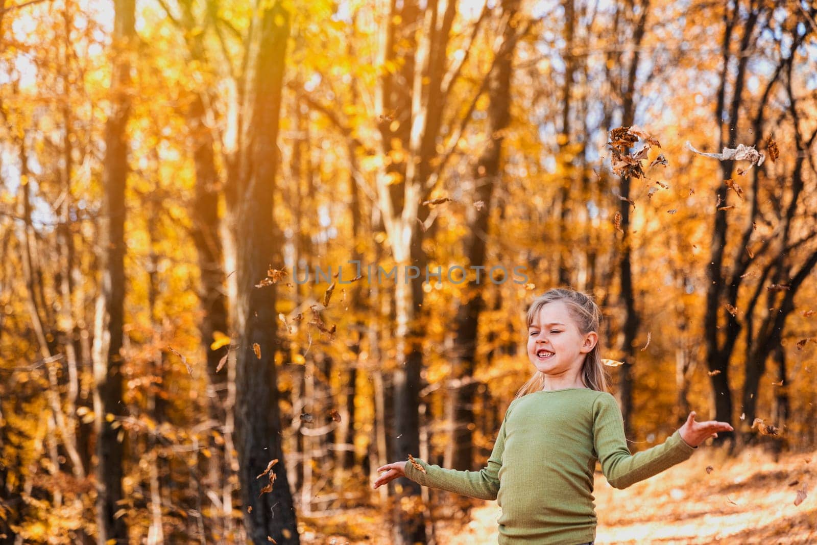 Small happy smiling little caucasian child girl playing with yellow leaves in autumn forest or garden around trees. Kid throws leaves up and they are falling down. Colorful autumn, gold colors around