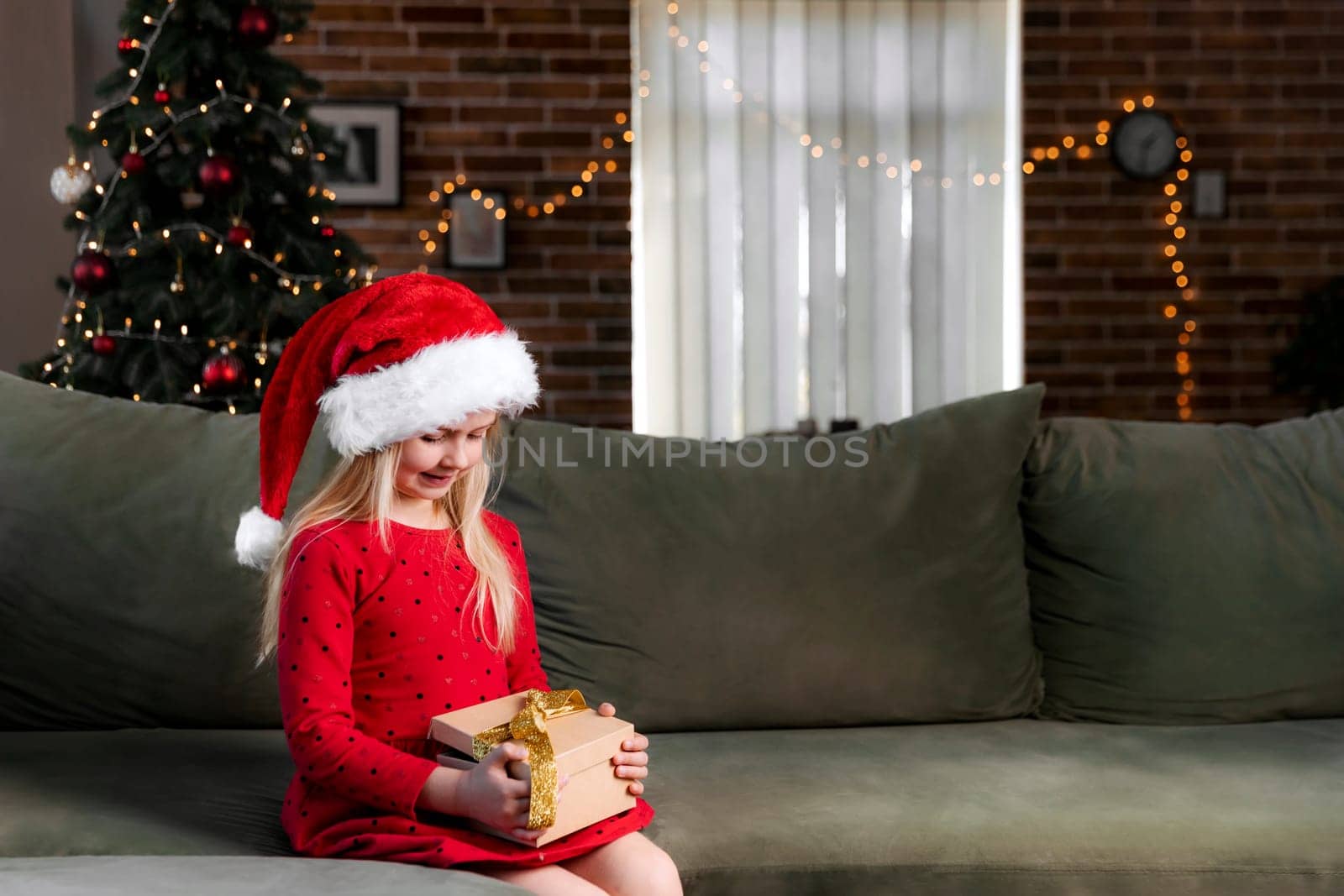 Small little smiling girl in red festive dress holding Christmas or New Year gift box present. Caucasian child dressed in Santa hat or cap sitting on green sofa in living room near decorated fir tree
