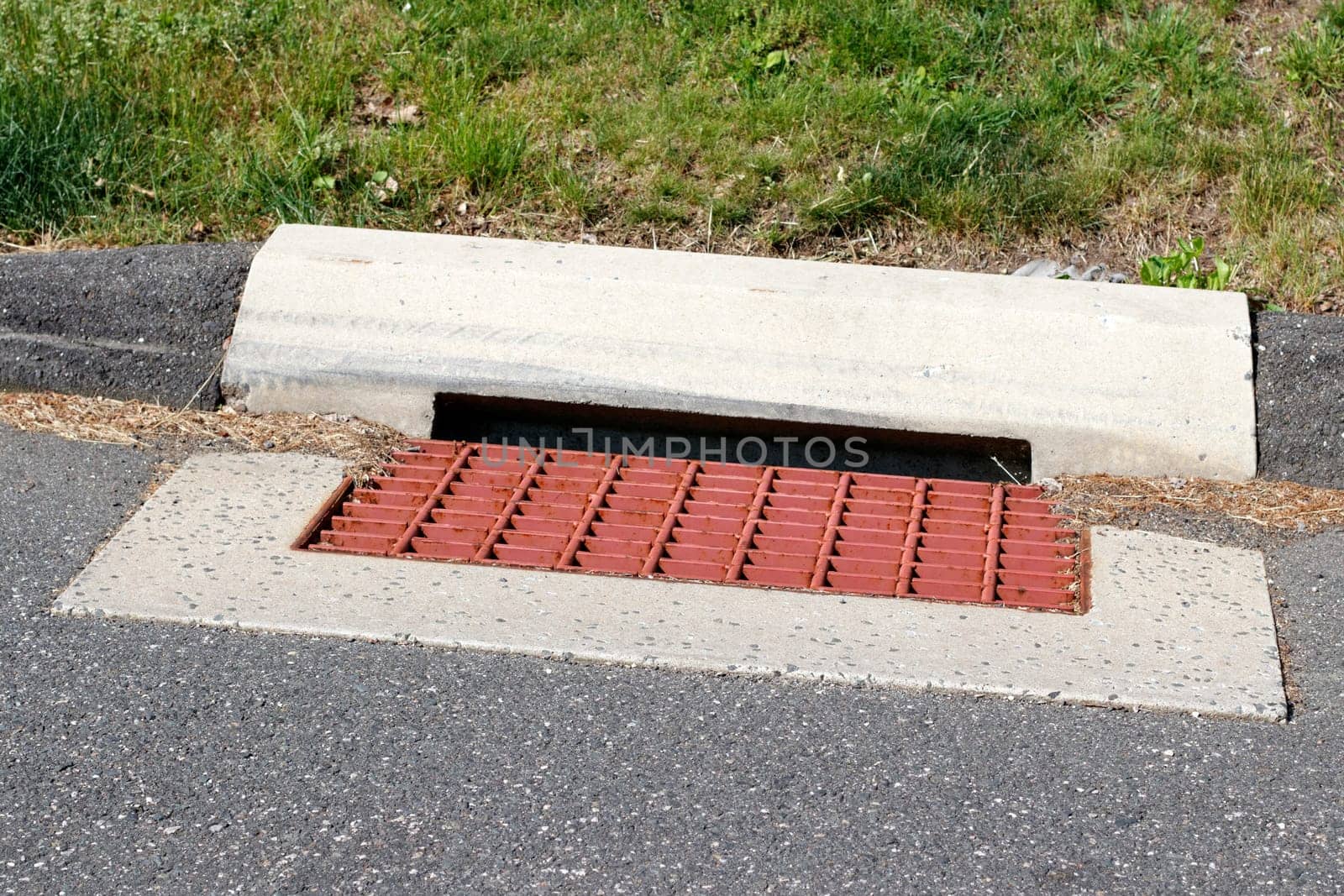Surface of a suburban street red painted metal storm water grill outdoors in the day with grass in back. Close-up of a red painted metal sewer grate surrounded by cement in a suburban curb and street