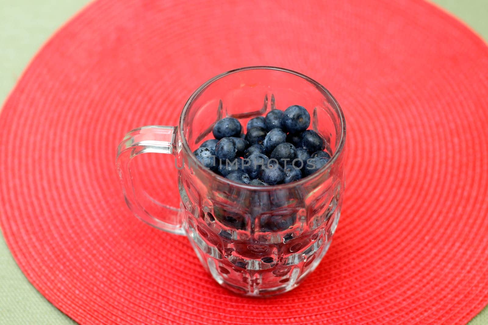 Large organic blueberry fruit in a clear glass cup on a round red place mat resting on a green table cloth. Many organic raw food blueberries filling a large clear glass mug on a red placemat closeup.
