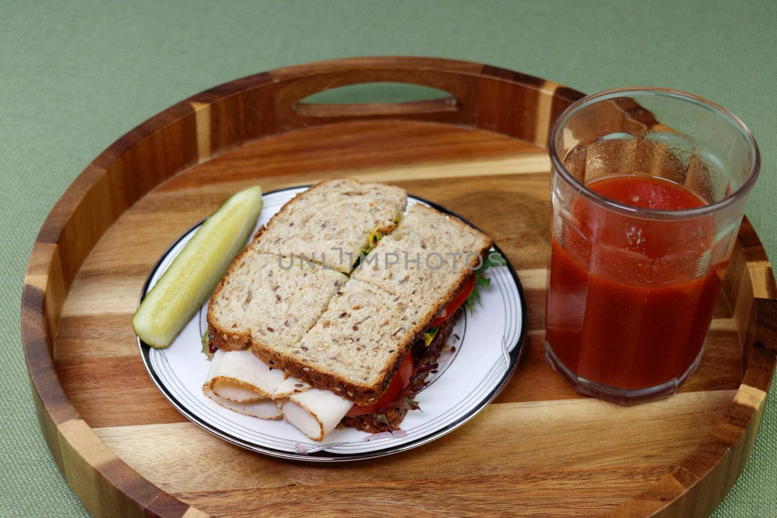 On a round wood tray on a plate is a turkey cold cut sandwich with tomato, lettuce and mustard near a vegetable beverage. Turkey and vegetable sandwich with a dill spear pickle and tomato drink meal.