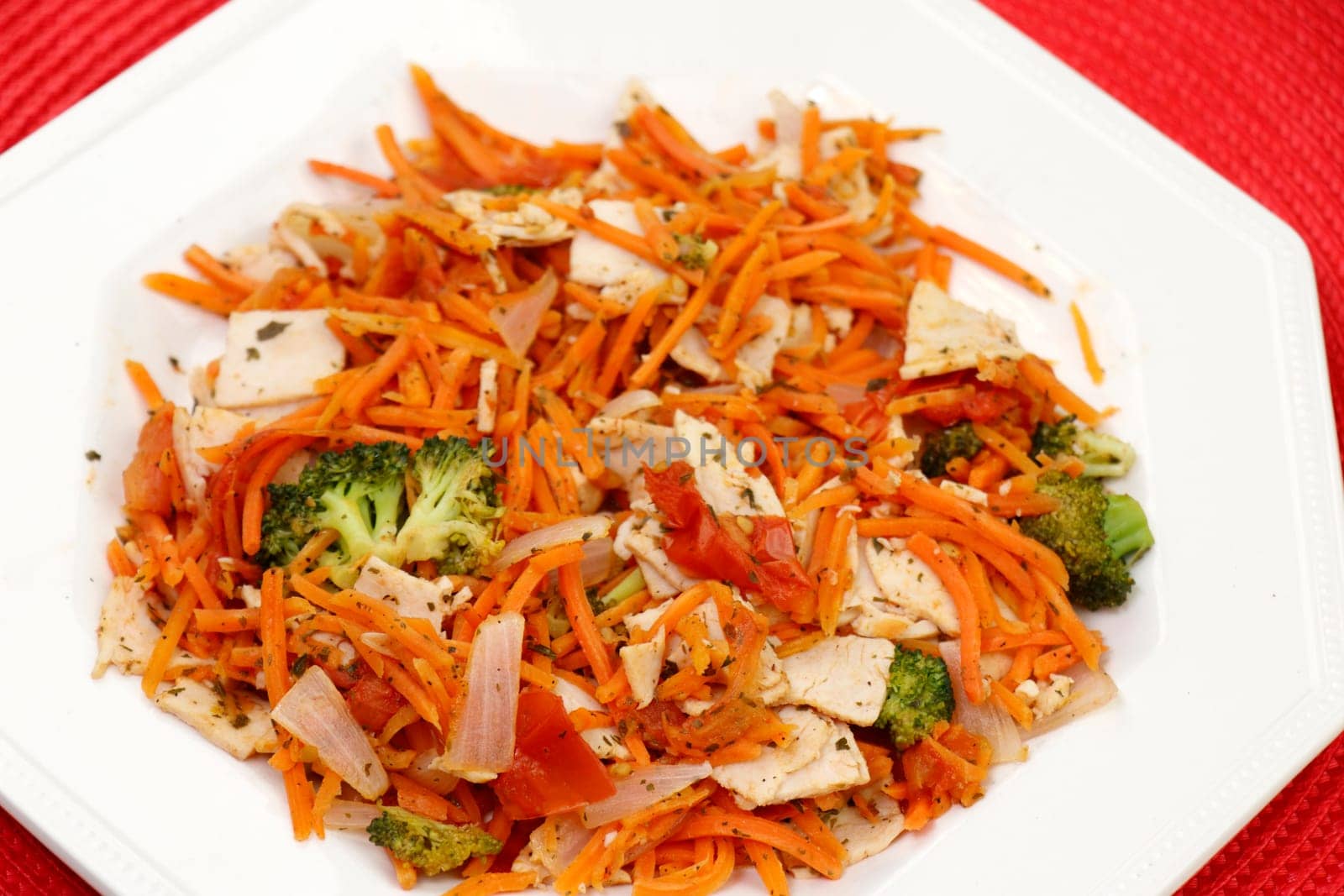Healthy lunch of cooked organic oven roasted turkey, carrot, broccoli, onion with parsley and black pepper. Close-up of cooked turkey and vegetables meal with seasonings on a plate on a red place mat.