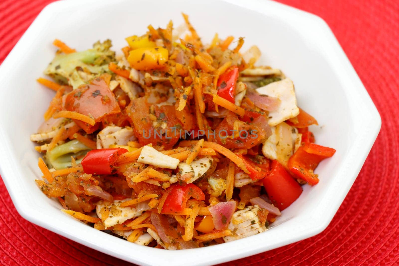In an octagon bowl is a prepared lunch of organic turkey meat and a variety of vegetables close-up on a red place mat. Cooked meal of turkey cold cuts sweet bell pepper and other vegetables and herbs