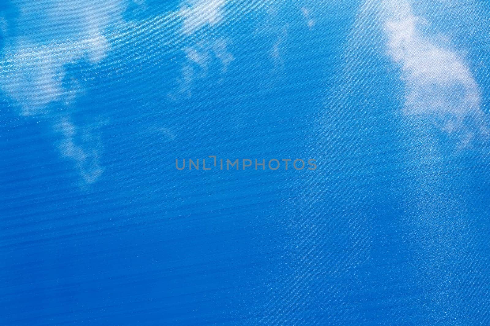Blue sunshade that you can see sunlight and white clouds above viewer that filters bright sunshine. Large flat blue sun shade textile structure that you can see through that reduces the sunshine below.