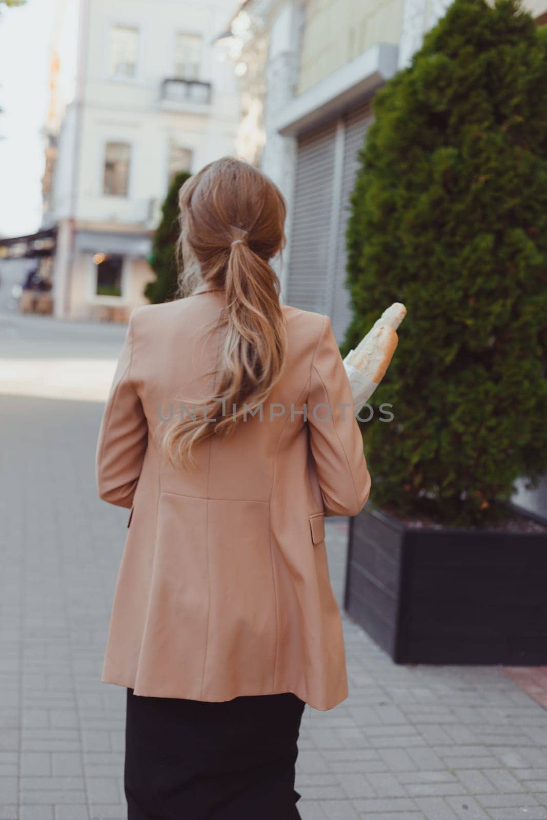 Business woman in a rush, carrying baguettes and takeaway hot beverage. Street portrait, back view.