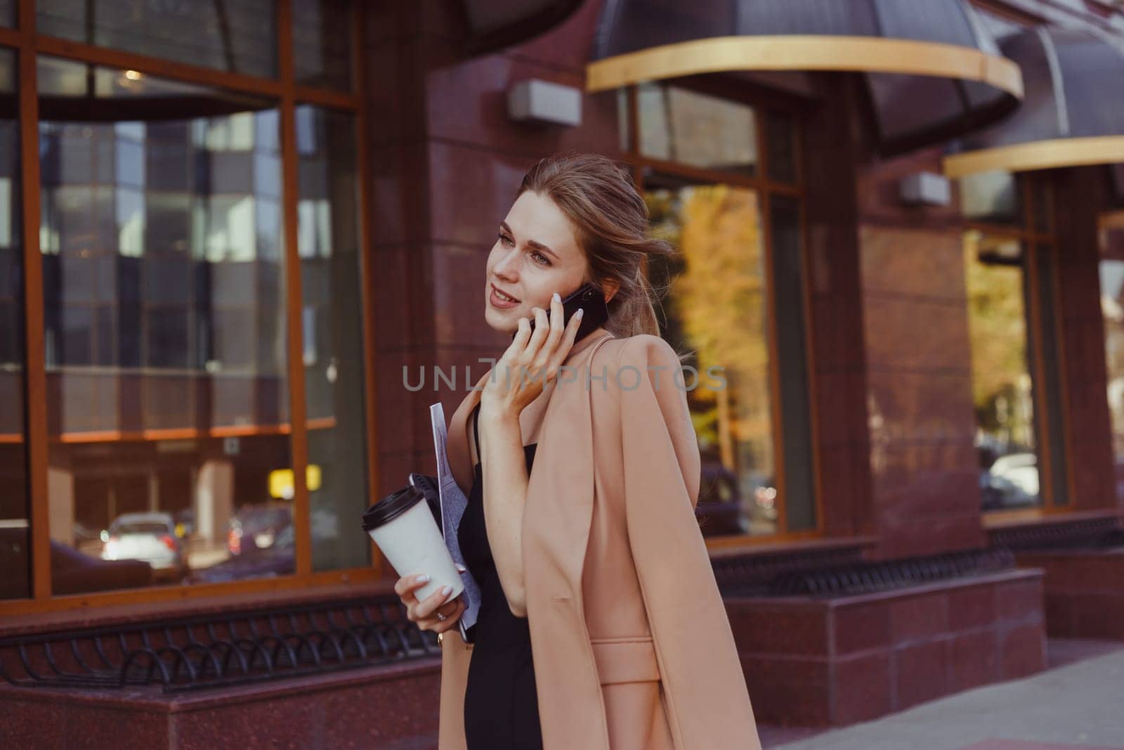 A busy woman with a newspaper, laptop and takeaway coffee in one hand is walking down the street and talking on the phone. Big city rhythm, rush