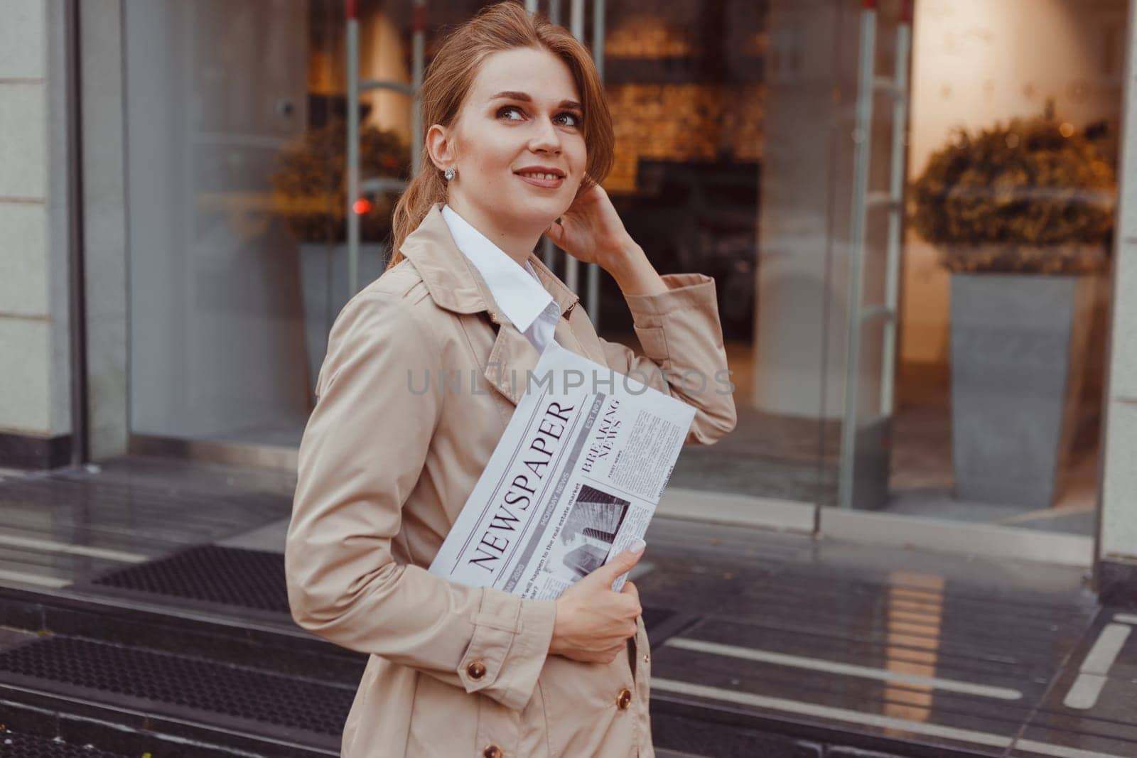 Beautiful girl in coat with newspaper and smiling while walking outdoors