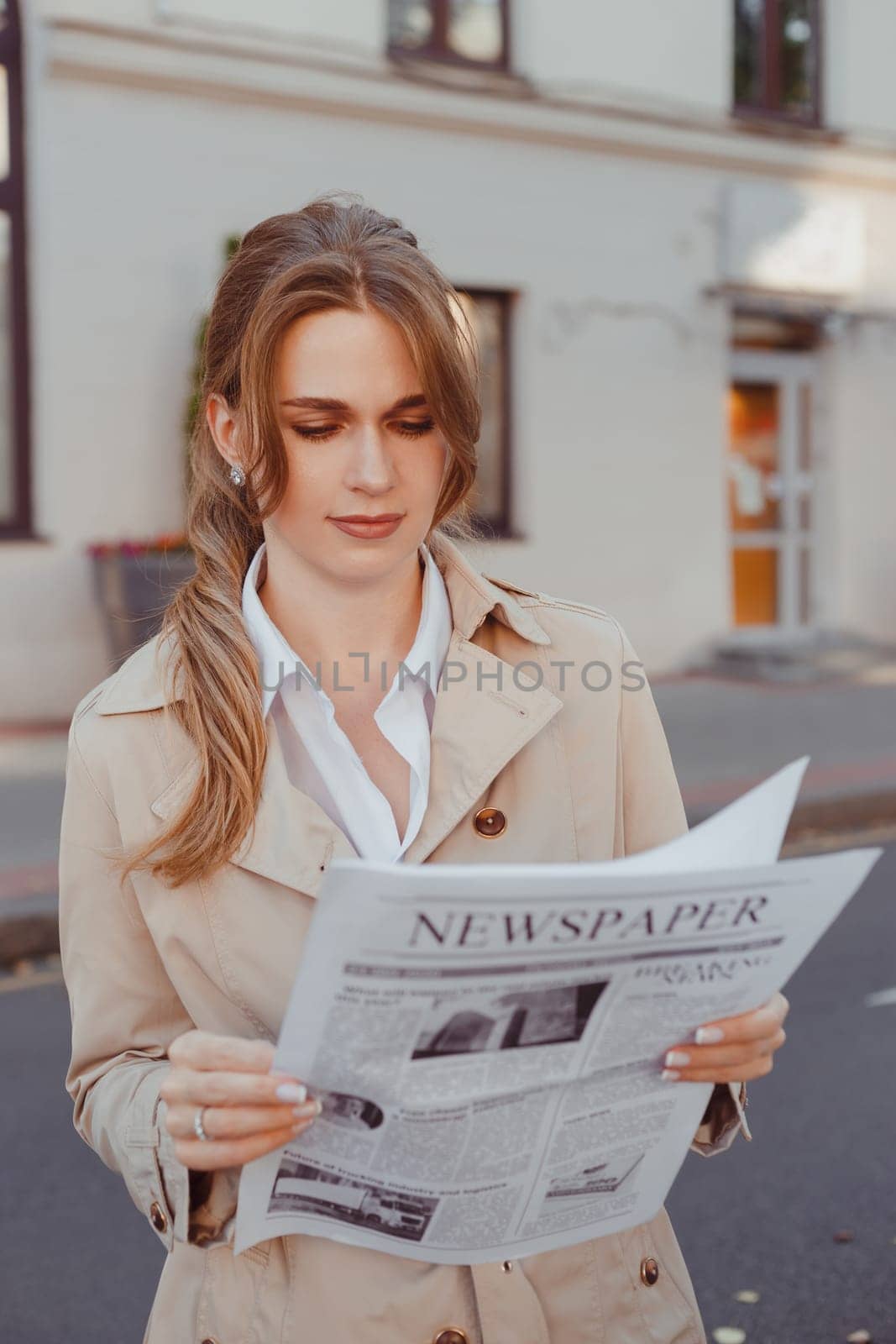 Attractive woman in beige coat reading newspaper standing outdoors.