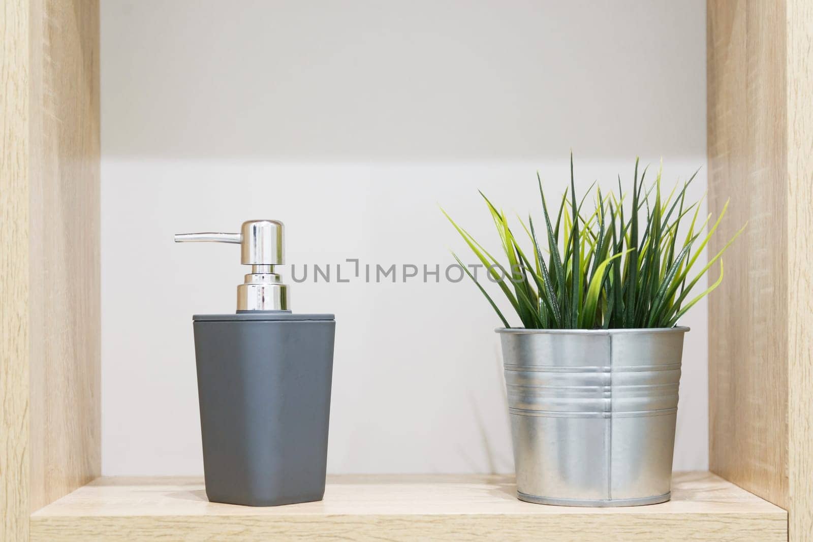 Soap dispensers and artificial houseplant in metal decorative pot on wooden shelf in bathroom. Interior details