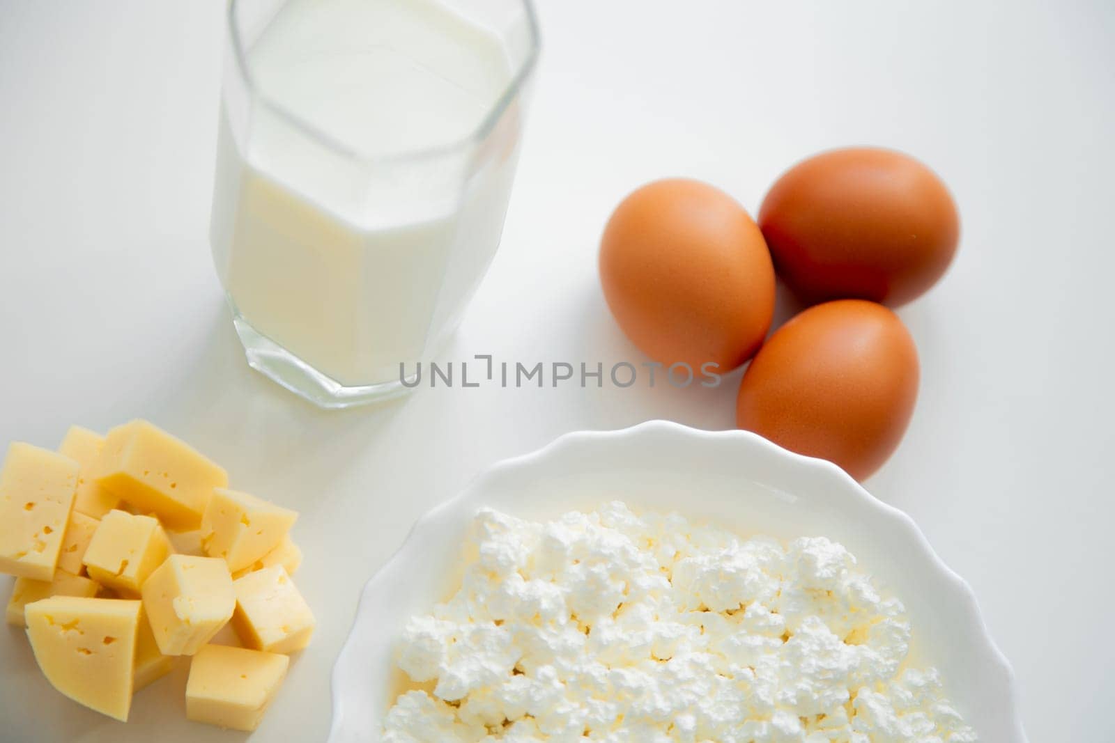 A white bowl with cottage cheese, eggs, diced cheese and a glass of milk stand on a white background. . High quality photo