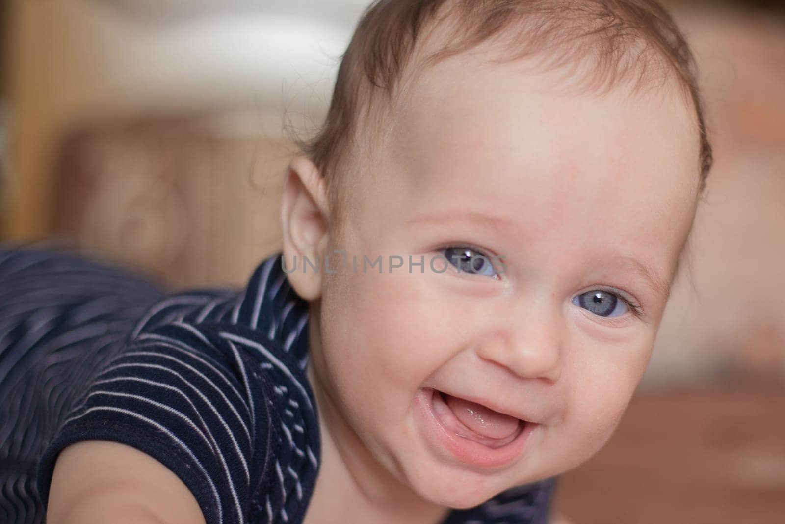 A toothless kid with beautiful blue eyes and a high forehead of Caucasoid appearance lies on his tummy and laughs into the camera in the blurred background. High quality photo