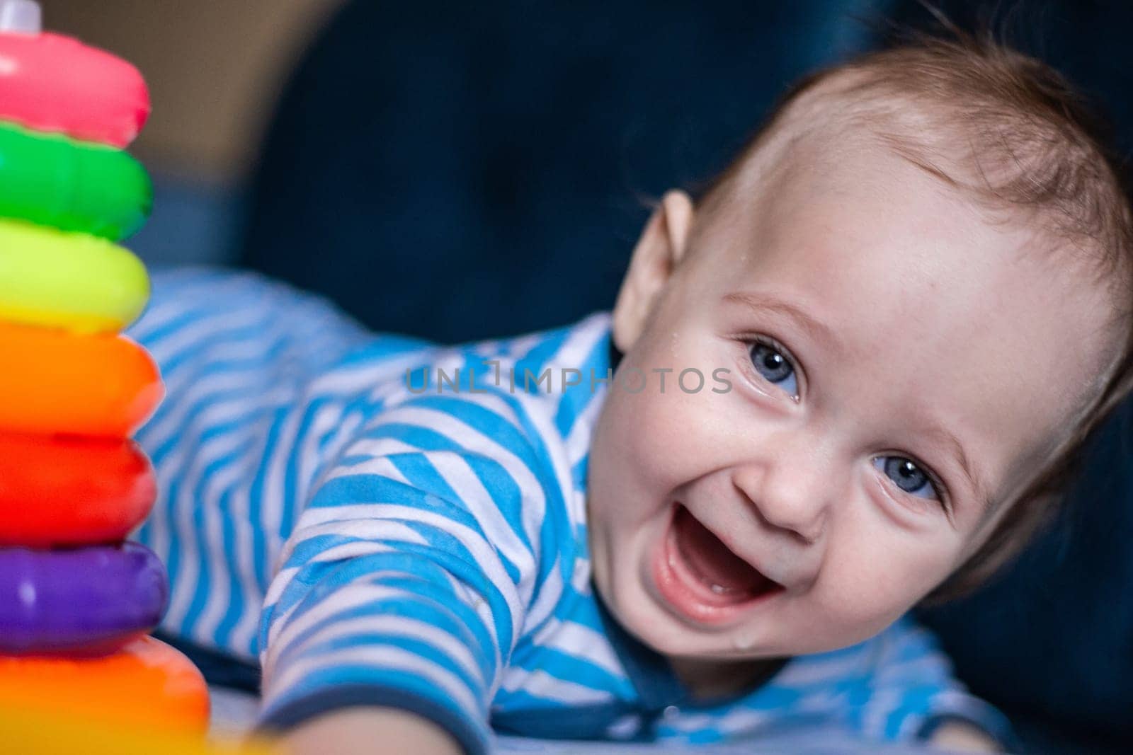 A little kid in a striped suit is lying on his tummy, next to him a children's pyramid and toys. High quality photo