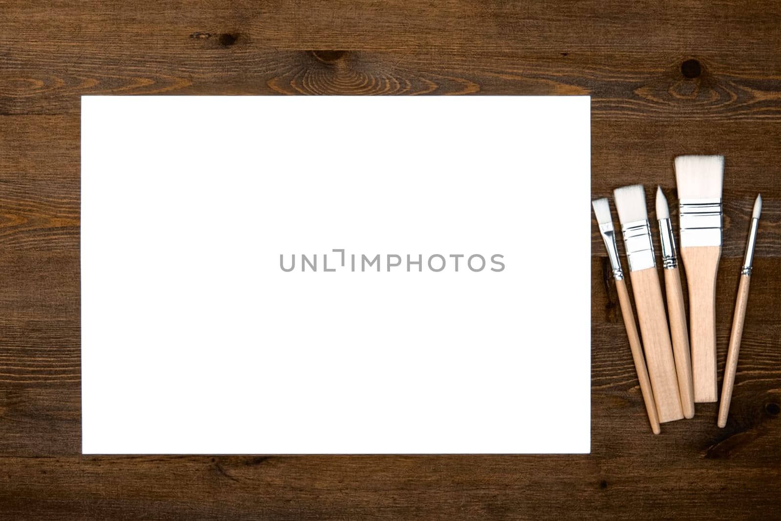 A clean white sheet and brushes on a wood textured background with space to copy. Layout, mockup space for text and drawing.