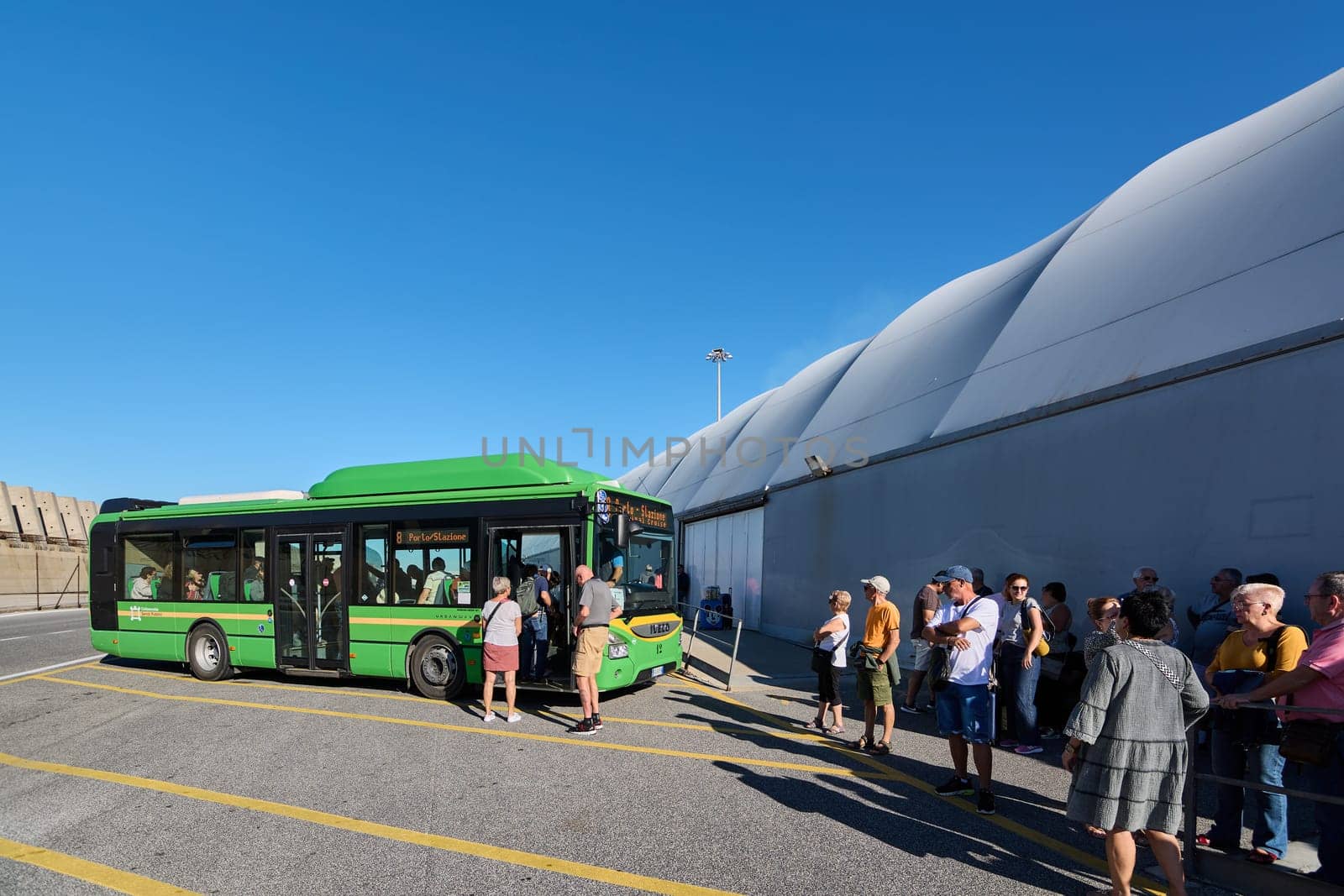 Civitavecchia. Italy - December 19, 2024: Passengers waiting in line next to a green bus at a stop on a sunny day, following the yellow line on the ground. Concept: Organized public transport.