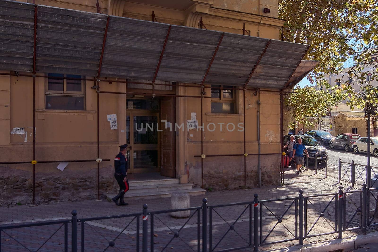 Civitavecchia, Italy - December 19, 2024: Old building with weathered facade and metal awning, with uniformed person walking and cars parked along the street. Concept: Urban history and everyday life.